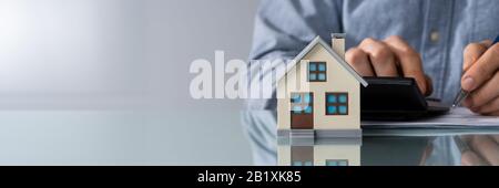 Close-up Of A Person Hand Doing Property Tax Calculation Using Calculator Of House Model Stock Photo