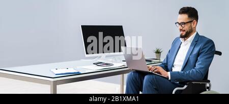 Full Length Of Young Handicapped Man Using Laptop While Sitting On Wheelchair At Home Stock Photo