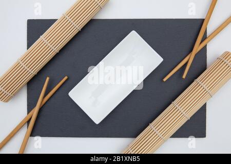 Rectangular slate plate with chopsticks, ceramic plate, bamboo mat for sushi on the white table. Stock Photo