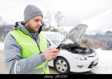Man using smart phone and calling road assistance emergency service. Broken car in the background. Winter scene. Stock Photo