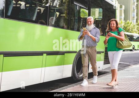 Happy senior couple using smartphones at bus station - 60's age people having fun with technology trends - Transport and joyful elderly concept - Focu Stock Photo