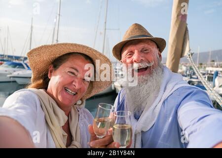 Happy senior couple doing selfie, cheering with champagne on a sailboat during anniversary vacation - Joyful elderly lifestyle, love, travel and holid