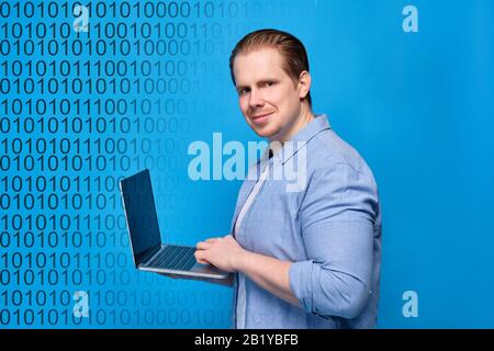 A man in casual clothes on a blue background looks at the camera and programs on a laptop. Binary code abstract. Close up. Stock Photo