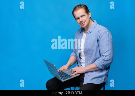 A man in casual clothes on a blue background looks at the camera sitting with a laptop. Close up. Stock Photo