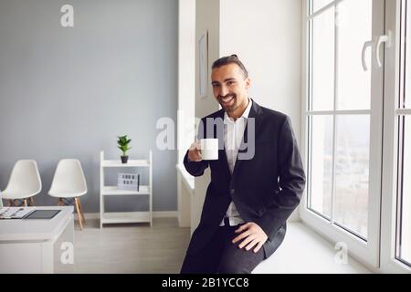 A man in a black jacket with a coffee mug in his hand is smiling while standing by the window in the office Stock Photo