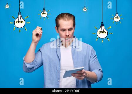 A man in casual clothes on a blue background holds a notebook and a pen wants to write down an idea. Abstract light bulbs burn on the background. Clos Stock Photo
