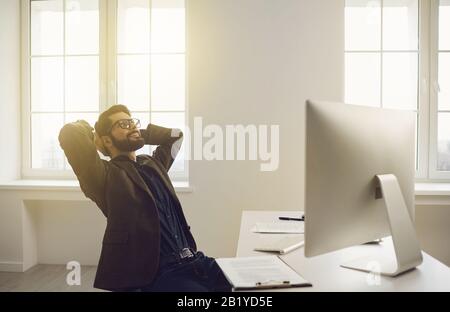 Businessman in glasses hands behind head contented dreams happy smiling sitting at a table in the office. Stock Photo