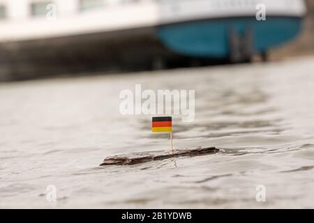 German Flag sticking in a piece of wood on a river with a big ship in the background Stock Photo
