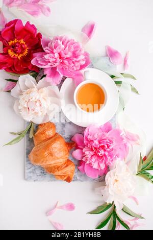 Breakfast for Valentines day with cup of tea, peony flowers and croissant on gray murble table and white background from above, flat lay, wedding, bir Stock Photo