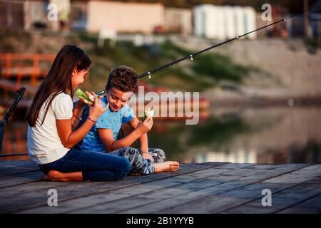 Two young cute little friends, boy and girl talking, eating sandwiches and fishing on a lake in a sunny summer day. Kids are playing. Friendship. Stock Photo