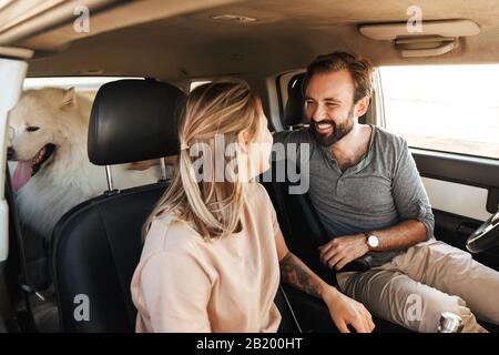 Image of happy young amazing loving couple with dog samoyed outdoors at the beach in car.