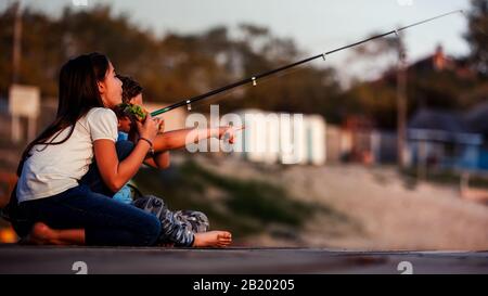 Two young cute little friends, boy and girl talkiing,eating sandwiches and fishing on a lake in a sunny summer day. Kids are playing and pointing with Stock Photo