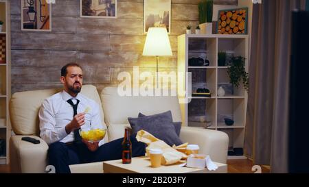 Businessman sitting on couch eating chips eating television. Zoom in shot. Stock Photo