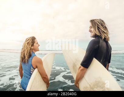 Surfers couple running together with surfboards on ocean at sunset time - Sporty fit friends having fun surfing on high waves Stock Photo