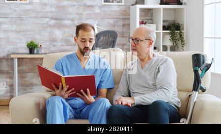 Social worker reading a book to an old disabled man in nursing home. Stock Photo
