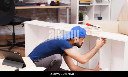 Attractive handyman assembles new furniture for home owners. Worker reading instructions from tablet computer. Stock Photo
