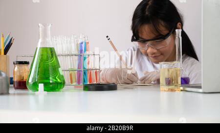 Photo of young adorable girl in safety glasses and gloves writing a experiment result while doing a scientist experiment at the modern laboratory with Stock Photo