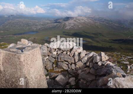 A view from the top of the Quinag mountain range, Scotland with the trig point, stone wind shelter, mountain tops and sky Stock Photo