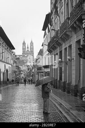 A man standing in the rain on a street in Quito, holding an umbrella with the Basilica raised high in the distance Stock Photo