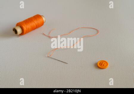 Button, spool of thread and needle with thread. Orange sewing accessories. Clothing repair. Side view. Selective focus. Stock Photo