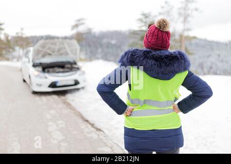 Rear view of stressed woman and broken car with opened hood in the background. Road trip problems and assistance concepts. Stock Photo