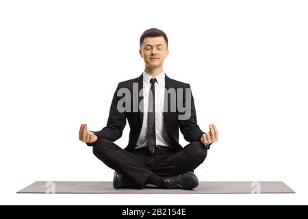 Young man in a black suit with closed eyes sitting with crossed legs and making a meditation pose on a mat isolated on white background Stock Photo