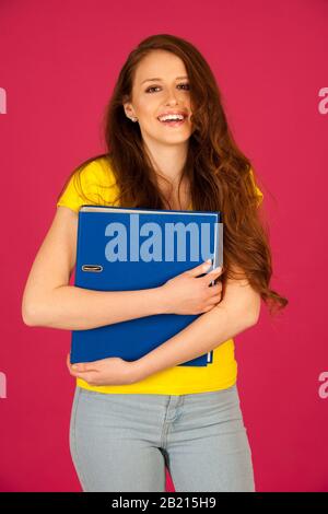 attractive young student in yellow t shirt holding red folder over pink background Stock Photo