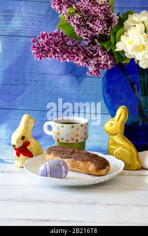 A light breakfast of a chocolate eclair and coffee, is garnished with colorful easter eggs and gold foil wrapped Chocolate bunnies
