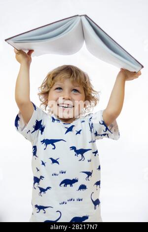Little boy between 4 and 5 years old holding a book over his head while smiling standing against white background Stock Photo