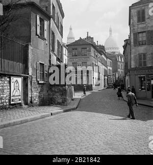 1950s, Paris, France, cobbled street in the Montmartre area of the Parisian city, famous for its street artists and church, Sacre Coeur. Picture shows Les Rue des Sales and the restaurant, 'Le Consulat' with the domes of the Sacre Coeur in the background. Stock Photo