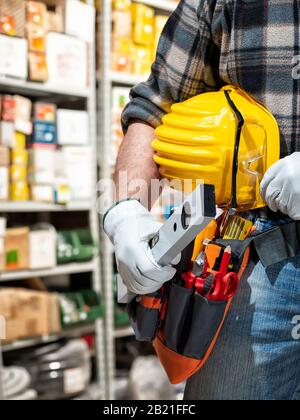 Electrician in the electrical parts store holds the level in hand, helmet with protective goggles. Construction industry, electrical system. Stock Photo
