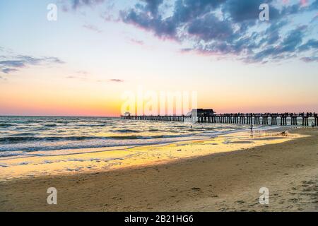 Naples, Florida yellow sunset in gulf of Mexico with sun behind Pier wooden jetty on horizon and silhouette ocean waves wide angle view with sand, peo Stock Photo