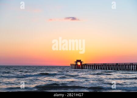 Naples, Florida vibrant red yellow sunset in gulf of Mexico with sun inside framing Pier wooden jetty on horizon and dark silhouette ocean waves wide Stock Photo