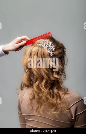 Wave curls hairstyle. Hairdresser making hairstyle to blond hair woman with long hair using comb on gray background. Professional hairdressing Stock Photo