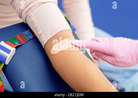 Doctor in rubber protective gloves putting an adhesive bandage on young woman's arm vein after blood test or injection of vaccine. Medical, pharmacy and healthcare concept. Stock Photo