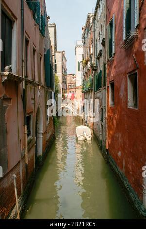 A small side canal with buildings, boat, and bridge. Laundry can be seen drying in the distance. Venice, Italy. Stock Photo
