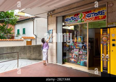 A female tourist reaches high up on a postcard display rack outside a colorful shop in Ventimiglia, Italy. Stock Photo