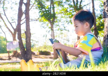 Adorable asian boy sweet casual sitting on green grass use tablet computer learnning nature tree in city park Stock Photo