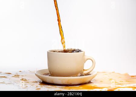 Coffee pouring into a white cup isolated against a plain white background, splashing in all directions creating a mess on the white surface Stock Photo