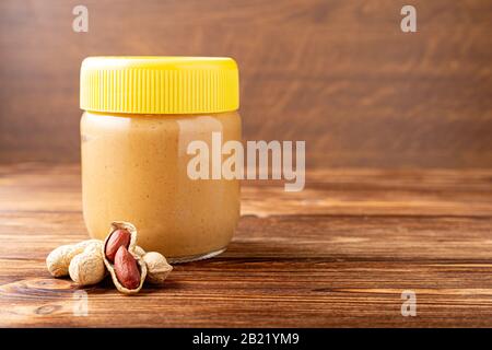 Creamy peanut paste in glass jar with yellow cap and peanuts in the peel scattered on the brown wooden table with copy space for cooking breakfast Stock Photo