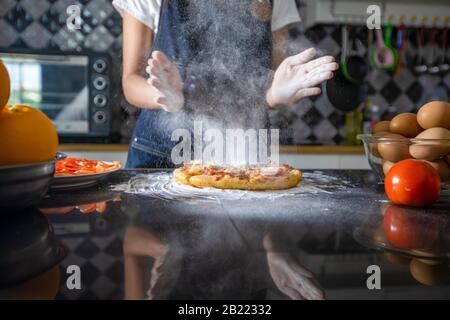 Asian women preparing a pizza, knead the dough and puts ingredients on kitchen table Stock Photo
