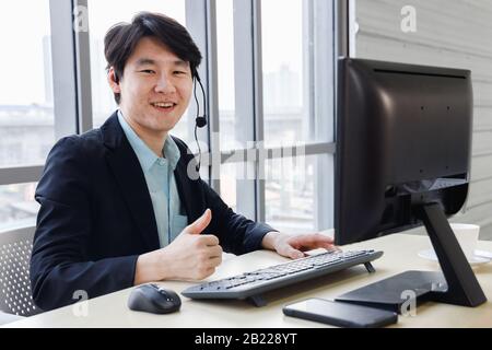 Smiling confident Asian male call center operator enjoy his work at office and perform thumb up gesture. Stock Photo