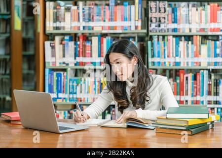 Asian young Student in casual suit doing homework and using technology laptop in library of university or colleage with various book and stationary ov Stock Photo