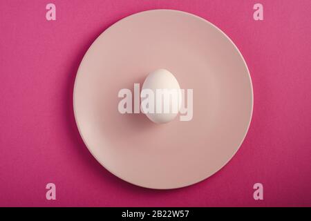 White egg in pink plate on purple plain minimal background, top view, happy Easter day Stock Photo
