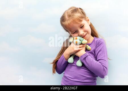 Little girl, happy child holding her stuffed cuddle rag toy close to her face smiling cheerful preschooler, hugging a small toy, clouds background Stock Photo