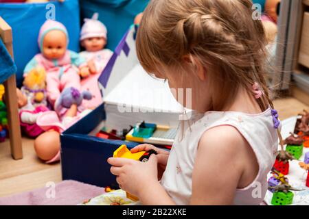 Little clever girl playing with building blocks construction car toy by herself alone in her room, seen from behind, dolls, bricks and many various to Stock Photo