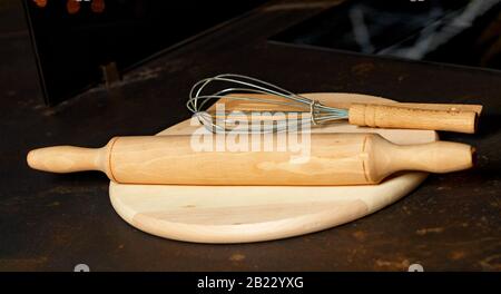 Wooden kitchen appliances rolling pin, whisk and spatula lie on a cutting board, brown table on a black background, selective focus, copy space. Stock Photo