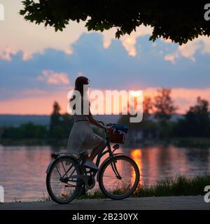 Silhouette young woman riding a bicycle, enjoying purple sunset at the lake, all colors of the sky, nature in the dusk, sun path, blurred background Stock Photo