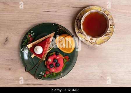 Two appetizing delicious red cakes and half of juicy orange on a glass plate with cup of tea nearby, top view Stock Photo