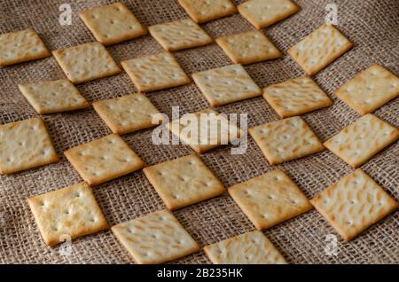 Square treats on burlap background. Geometric pattern of salted crackers. Repeating loop ornament. Selective focus. Close-up. Stock Photo